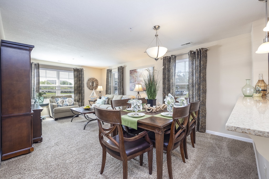 A contemporary living and dining room with a wooden dining table set for four, neutral-toned sofas, and large windows bringing in natural light. Decor includes plants and modern art.