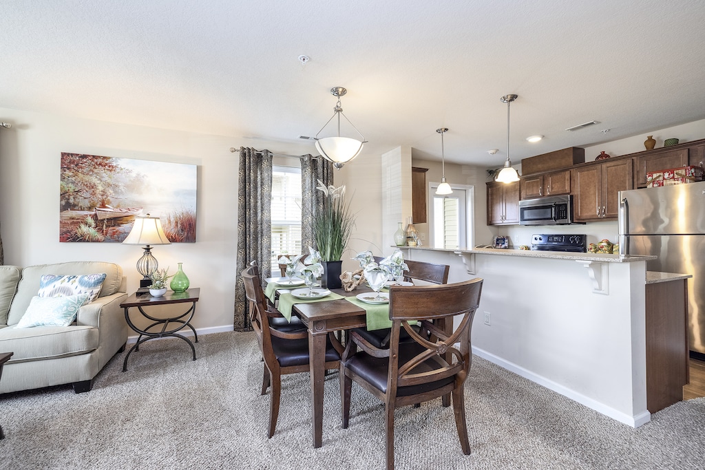 Dining area with a wooden table and chairs in an open-plan living space. The kitchen features stainless steel appliances. Decor includes plants, artwork, and pendant lights.