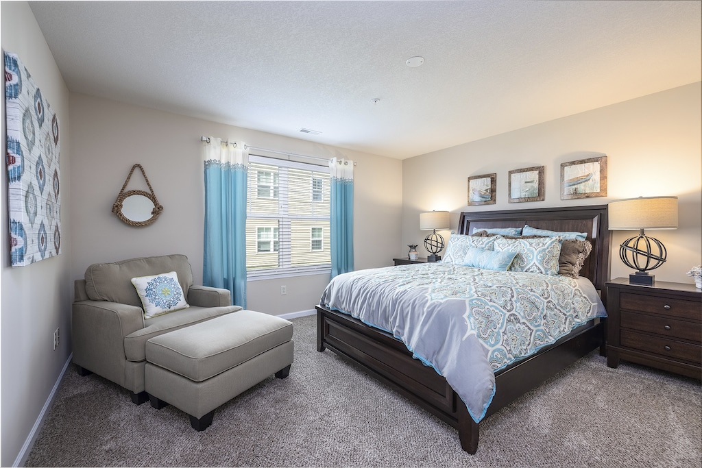 A bedroom with a bed, patterned bedding, two lamps on dark wood nightstands, a cushioned chair, and wall decor, including a mirror. A window with curtains lets in natural light.