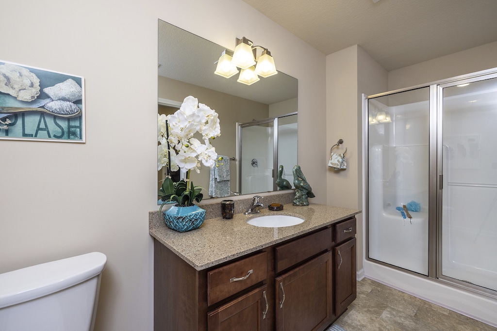 Bathroom with a large mirror, sink, and wooden cabinet. Orchid plant and toiletries on the counter. Shower with glass door on the right. Art depicting seashells on the wall.