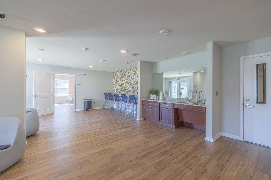 A modern room with wooden flooring, chairs lined against a wall, a countertop with a sink, and a colorful wall mural. Bright overhead lighting illuminates the space.