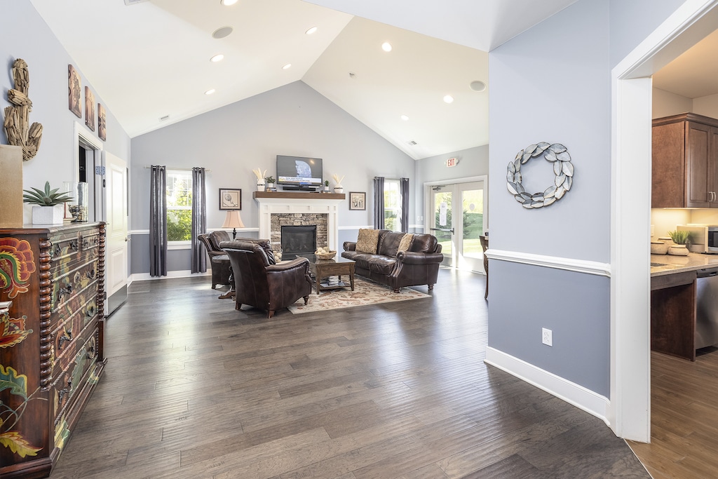 Spacious living room with vaulted ceiling, dark hardwood floors, leather furniture, and a stone fireplace. Natural light streams through large windows and a glass patio door.