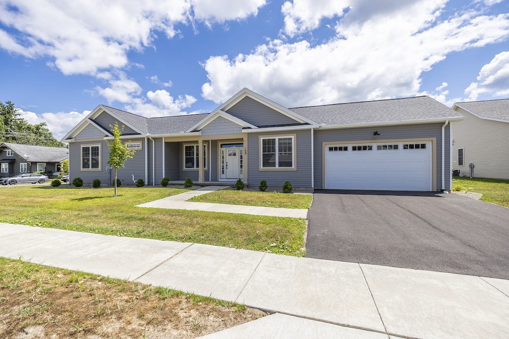 Single-story gray house with white trim, a neatly maintained front lawn, and a two-car garage. Sidewalk and driveway visible in the foreground, under a partly cloudy sky.