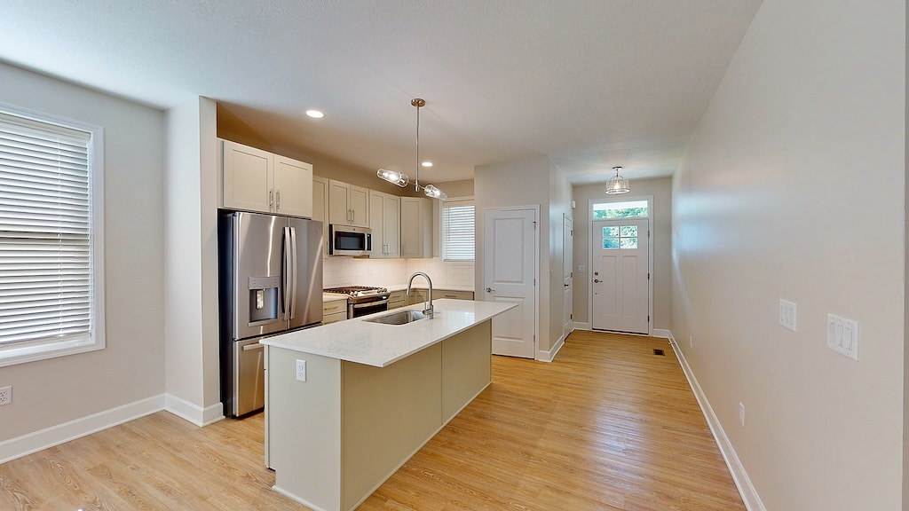 Modern kitchen with a center island, stainless steel appliances, and pendant lighting. Light wood flooring and white walls create a bright atmosphere. Entryway visible in the background.