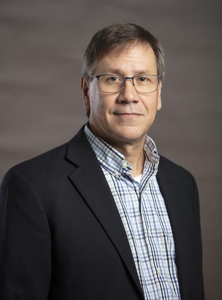 A man in a suit jacket and checkered shirt stands against a plain background, wearing glasses and looking at the camera.