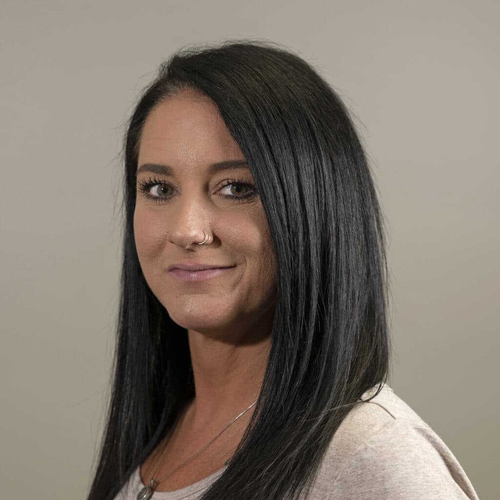 A woman with long black hair and a nose ring is looking at the camera against a plain background. She is wearing a light-colored top.