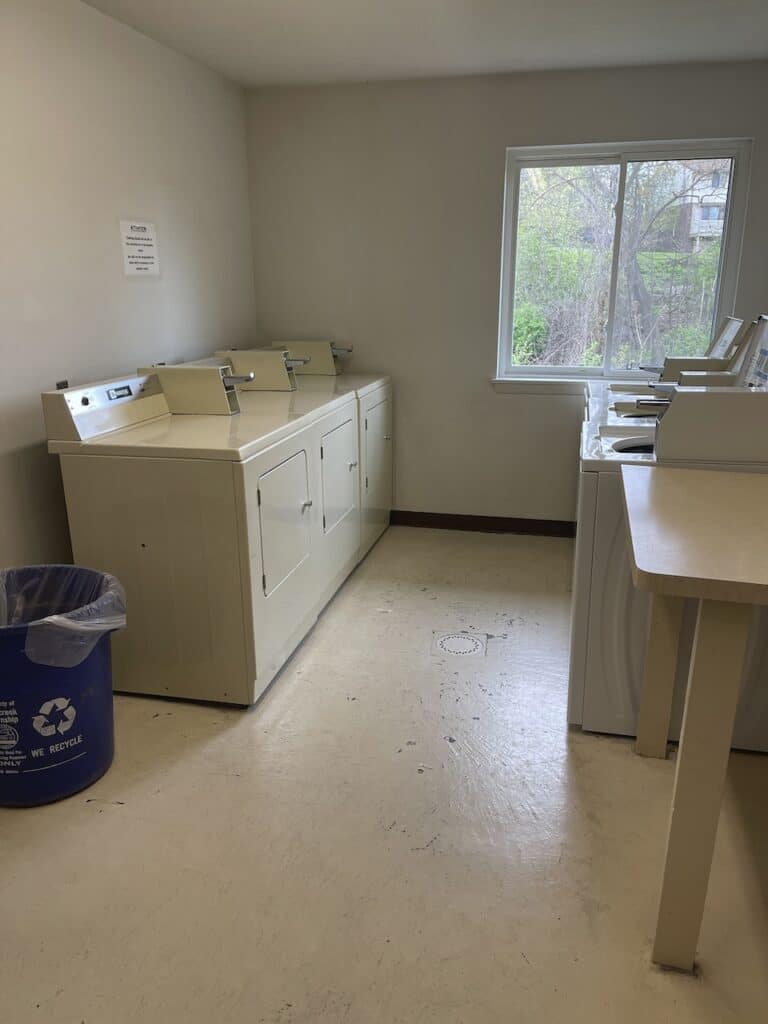 A small laundry room with two washing machines, a recycling bin, and a window overlooking trees.