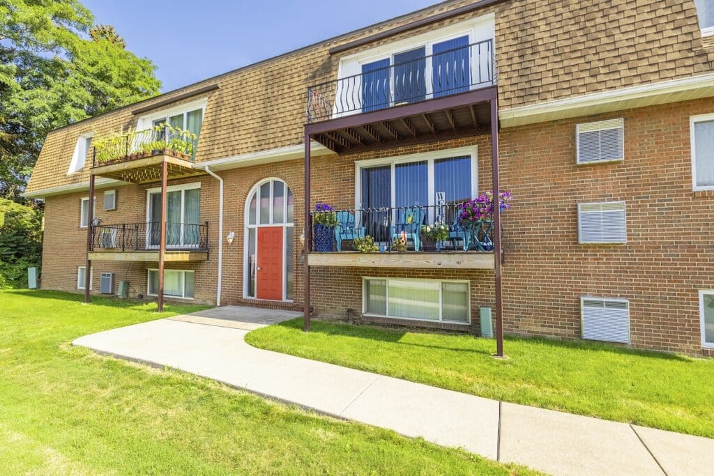 A brick apartment building with two balconies and a red door. The building has a shingled roof and a sidewalk leading up to the entrance. Grass and trees surround the area.