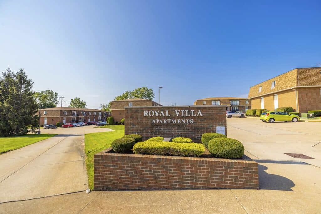 Entrance sign for Royal Villa Apartments, surrounded by shrubs, with brick apartment buildings and parked cars in the background.