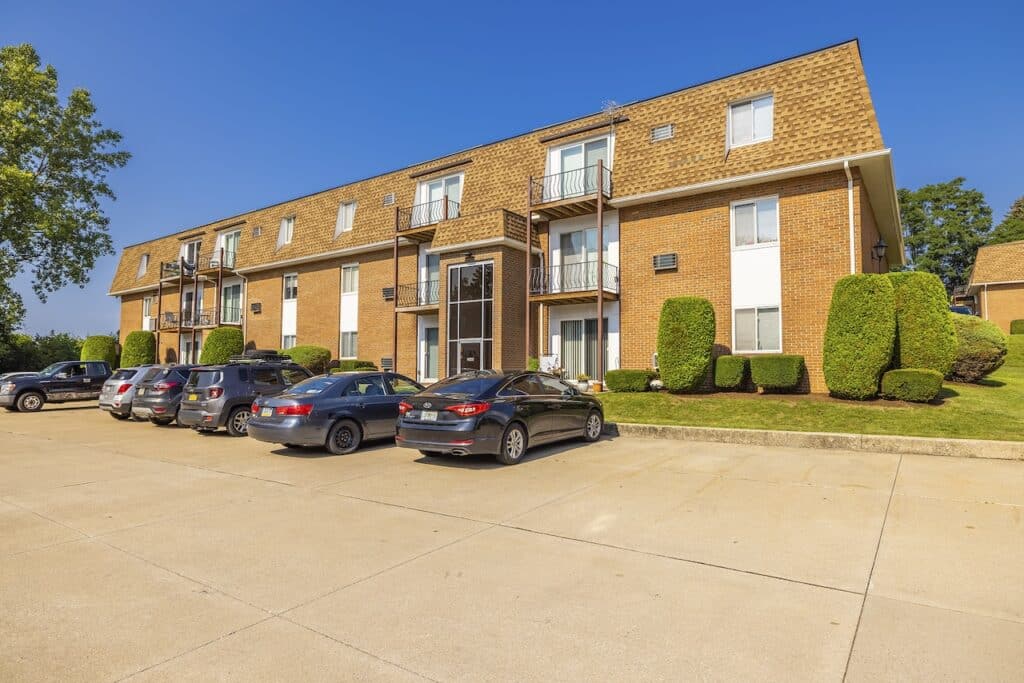 A three-story brick apartment building with balconies and parked cars in front, set against a clear blue sky. Trimmed bushes and grass are visible in the foreground.