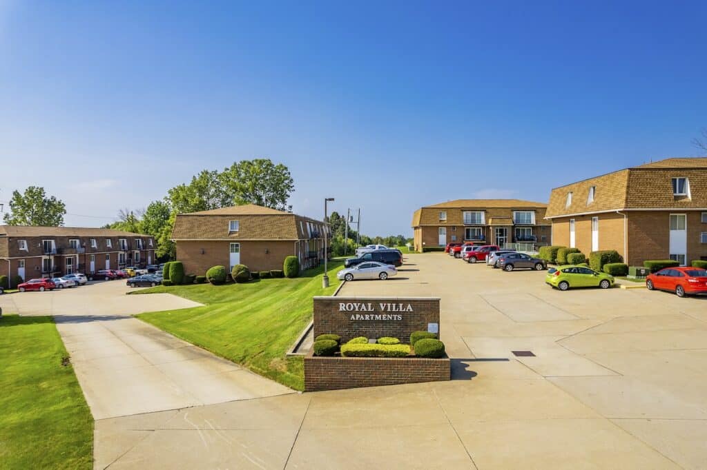A well-maintained apartment complex with multiple brown brick buildings, a central parking lot with parked cars, and green lawns under a clear blue sky.