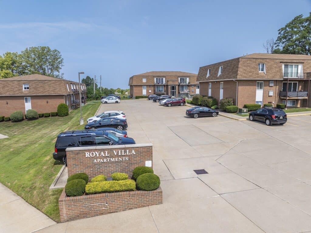 Entrance view of Royal Villa Apartments showing two brick buildings, a parking lot with cars, and a landscaped area with a sign.