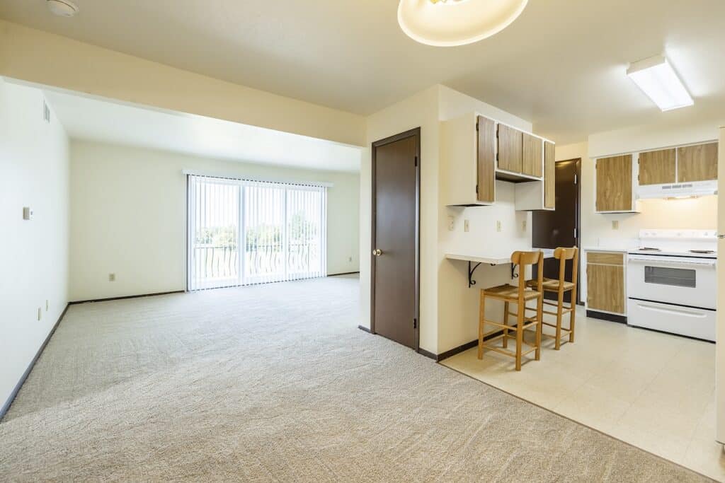 Open living space with carpeted flooring, adjoining a kitchen area with wood cabinetry and two bar stools. A sliding glass door leads to a balcony, allowing natural light to fill the room.