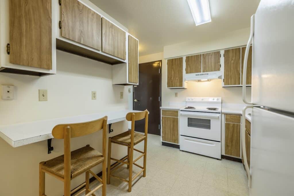 A kitchen with wooden cabinets, a white stove, fridge, and countertops, along with two wooden bar stools.