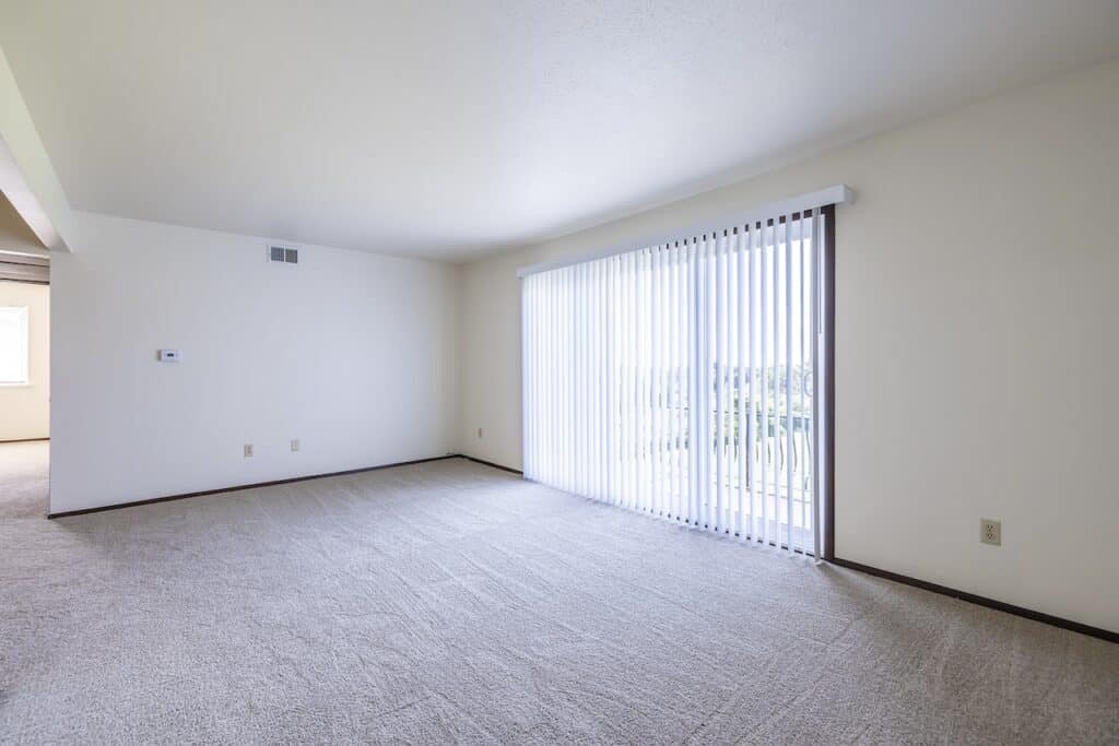 Empty room with beige carpet and white walls, featuring a large window with vertical blinds. Natural light fills the space.