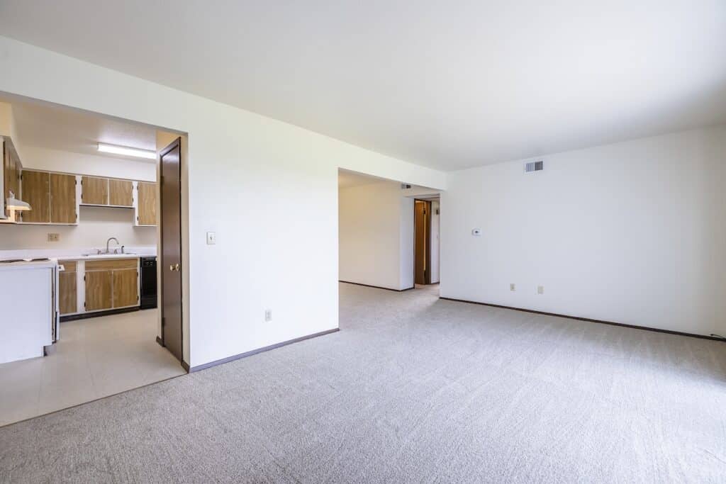 Open floor plan view of the living room with beige carpet and white walls, leading to a kitchen with wood cabinets and black appliances.