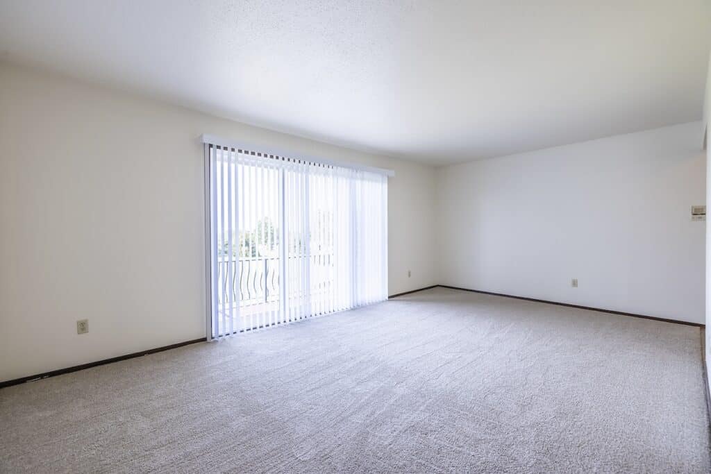 An empty room with beige carpet and white walls, featuring large sliding glass doors with vertical blinds leading to a balcony.