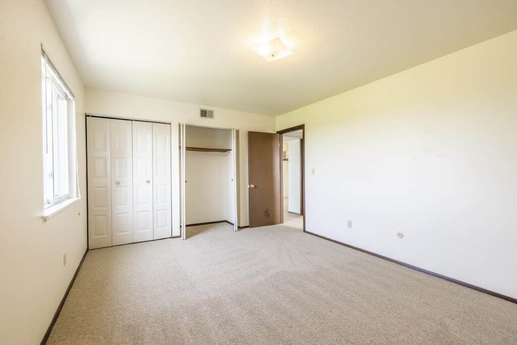 Empty room with beige carpet, white walls, a window on the left, and an open closet with double bi-fold doors. A light fixture is on the ceiling.