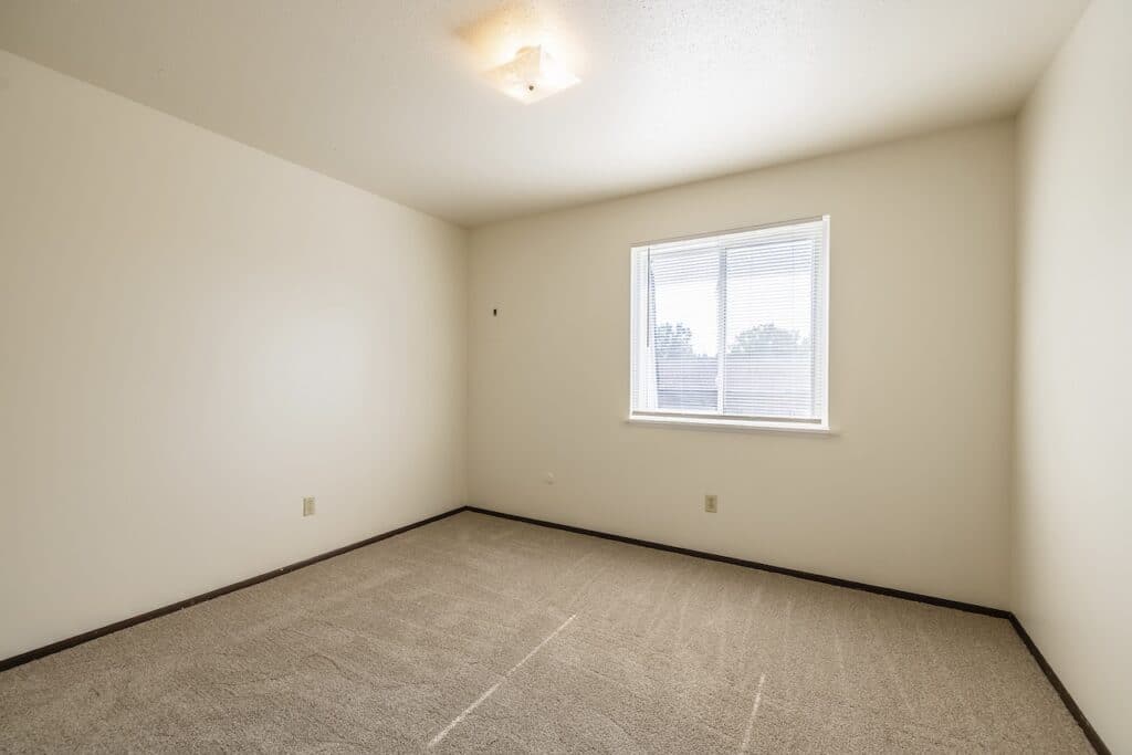 An empty room with beige carpeting, white walls, and a small window letting in natural light. A ceiling light fixture is centered above.