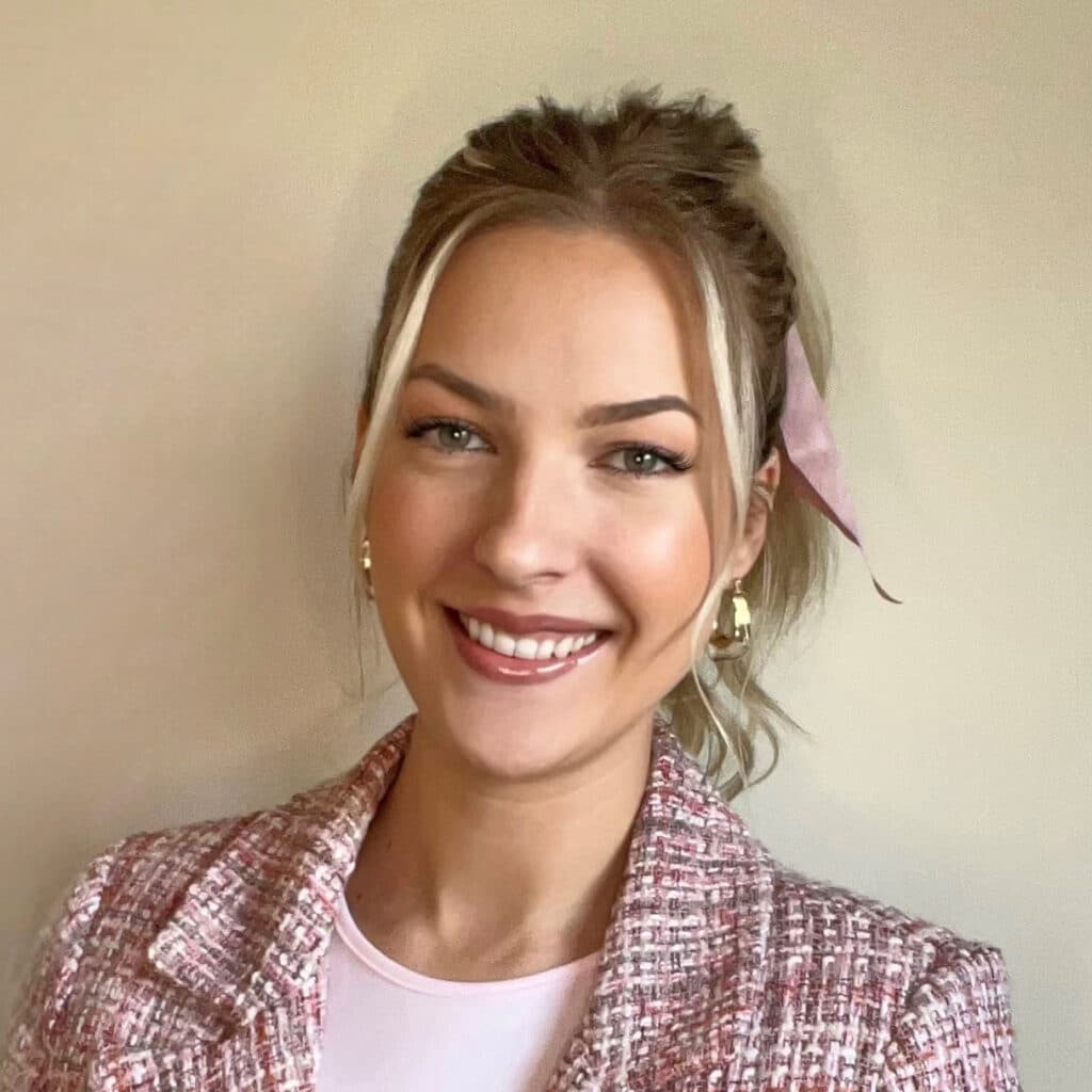 A person with blonde hair tied back and a pink ribbon smiles while wearing a pink and white checkered jacket.