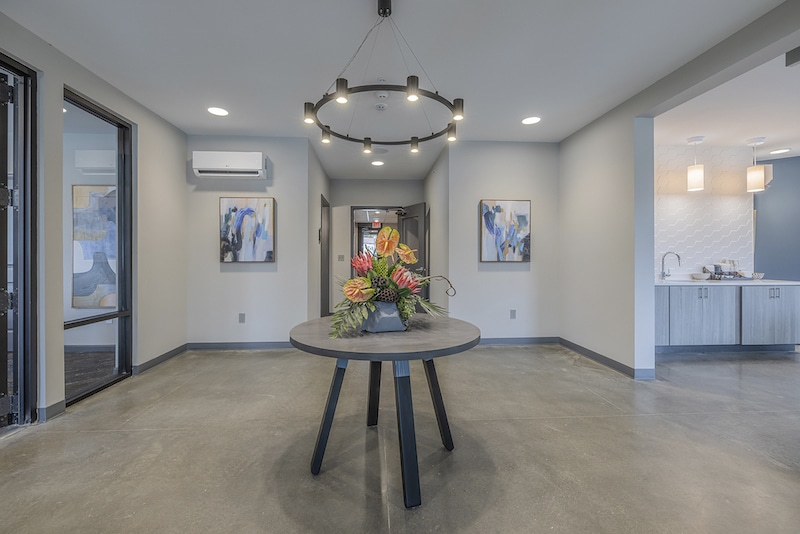 A modern foyer with a round table displaying a floral arrangement. Abstract paintings are on the walls, and a kitchen is visible on the right.