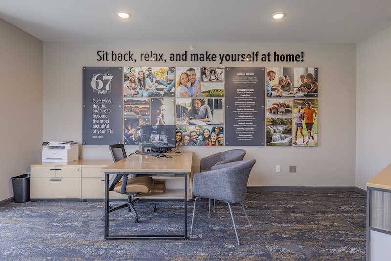 Office space with a wooden desk, chairs, and a printer. The wall displays photos and the text: "Sit back, relax, and make yourself at home!" along with a community message.