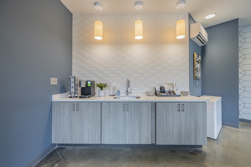A modern kitchenette with a hexagonal tile backsplash, sink, coffee machine, and overhead lighting. Cabinetry is light wood, with a small plant and decorative items on the counter.