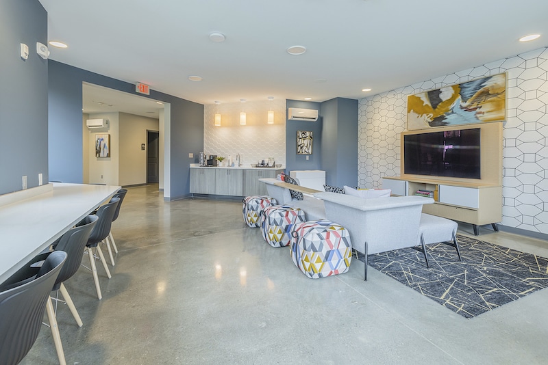 Modern lounge area with a long table and chairs on the left, a seating area with a sofa and colorful stools in the center, and a large TV on the right wall.