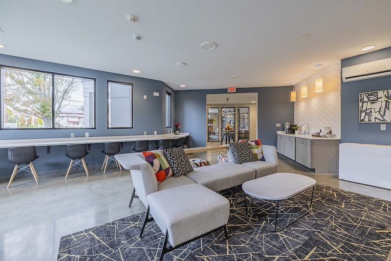 Modern lounge area with a gray sectional sofa, patterned rug, bar seating by the window, and a kitchenette. Large windows provide natural light, and decorative lighting fixtures hang from the ceiling.