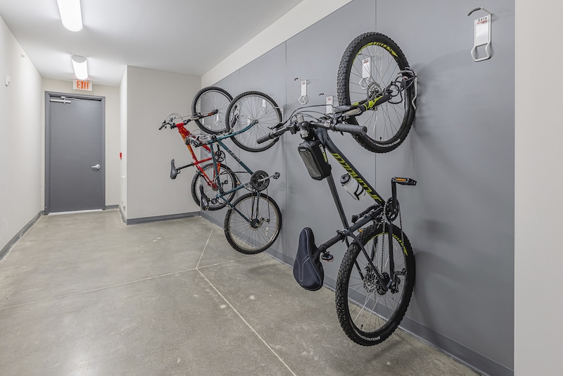 Three bicycles are vertically mounted on a gray wall in a clean, well-lit hallway with concrete floors and a closed gray door.