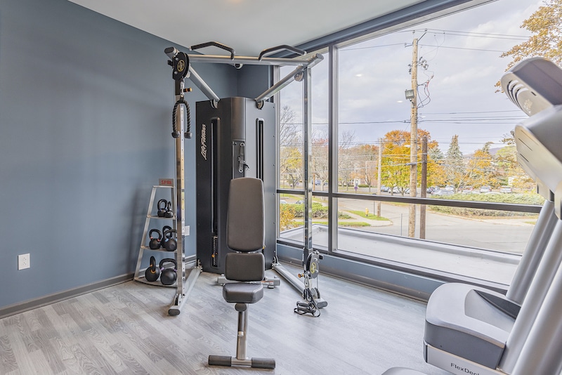 A gym room with exercise equipment including a multi-station machine, kettlebells on a rack, and a treadmill by large windows overlooking a parking lot.