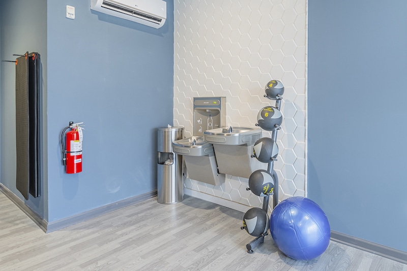 Gym corner with wall-mounted dual water fountain, trash can, fire extinguisher, five medicine balls on a rack, and a blue exercise ball on the floor against a blue wall.
