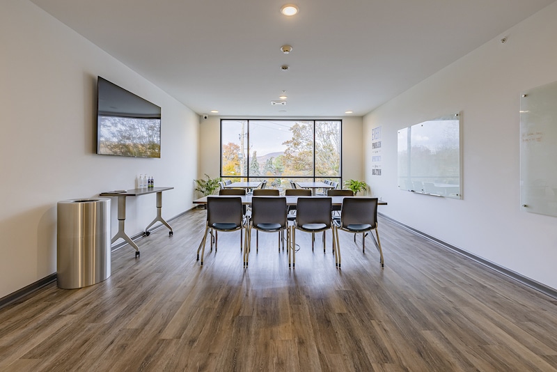 A conference room with a long table, black chairs, a large wall-mounted screen, whiteboards, and a window view of trees.
