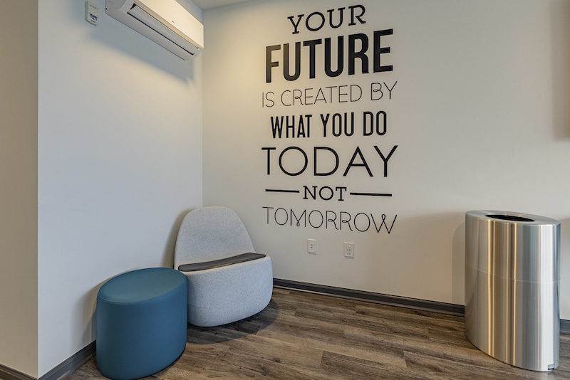 A modern room with a gray chair, blue ottoman, and silver trash can. Wall decor reads, "Your future is created by what you do today, not tomorrow." Air conditioning unit above.