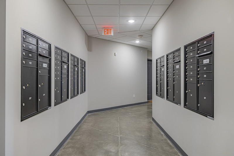 A hallway with two walls of numbered black mailboxes and a gray tile floor, featuring an illuminated exit sign on the ceiling.