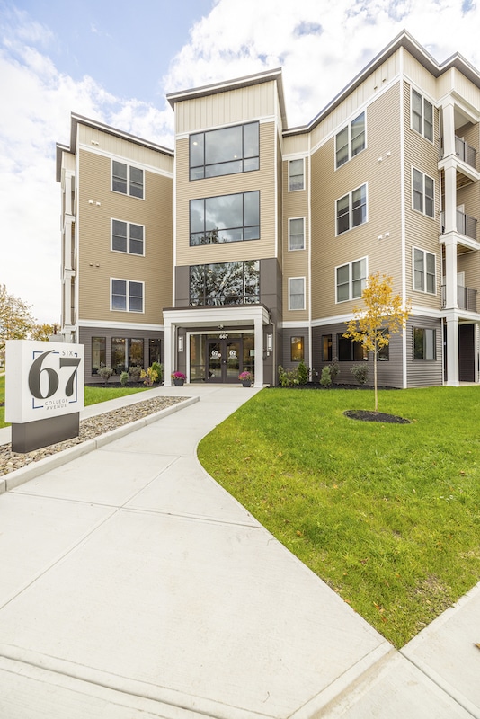 Four-story beige and gray building with large windows, lush green lawn, and a "67" sign in front, under a partly cloudy sky.