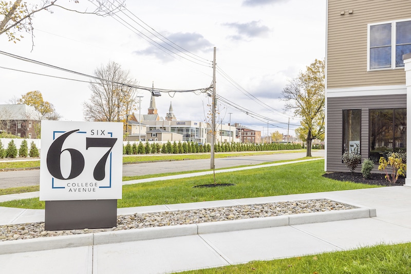 Sign reading "67 College Avenue" near a sidewalk, with trees and buildings in the background.
