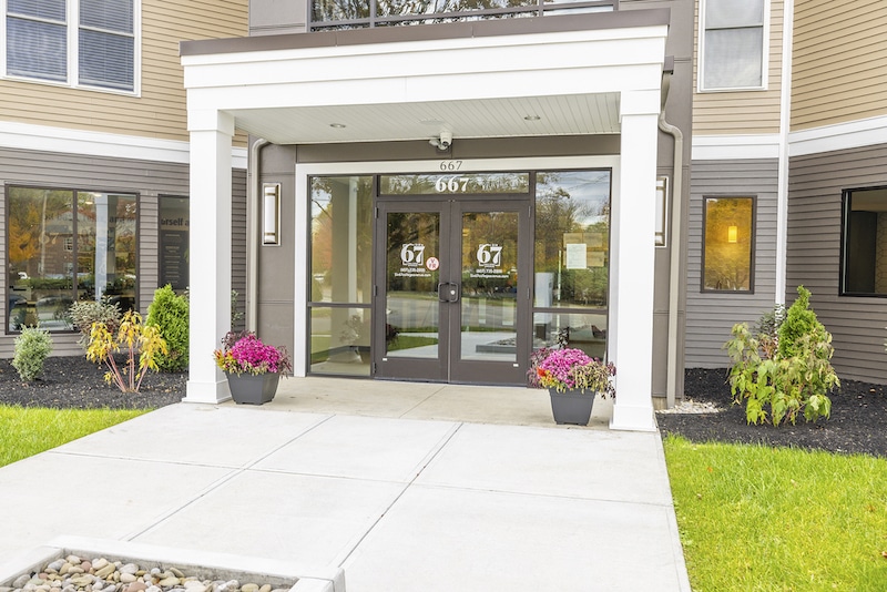 Entrance of a building with glass doors and sidelights, marked "67," featuring potted plants on each side, a sidewalk in front, and a reflective glass area above the entrance.