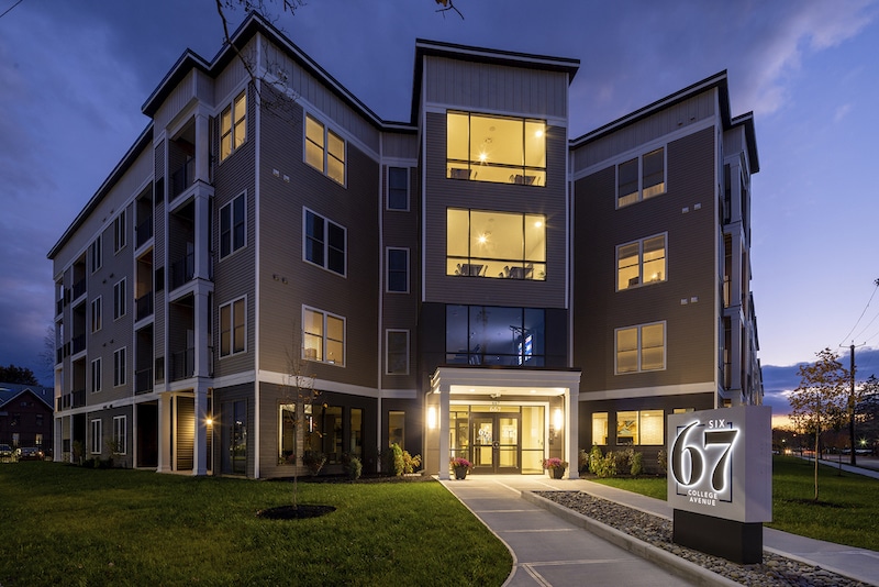 Four-story apartment building at dusk, illuminated with warm interior lights. In the foreground, a sign reads "Sixty67 Apartments." The sky is partly cloudy.