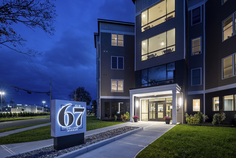 Exterior view of a modern apartment building at night with illuminated signage reading "Six67 College Avenue.