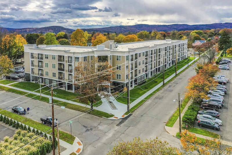Aerial view of a four-story apartment building with a parking lot and surrounding trees, under a cloudy sky.