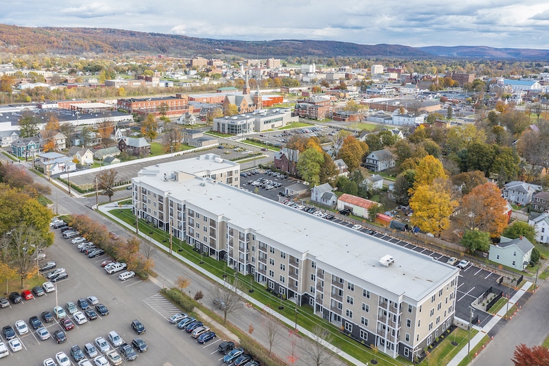 Aerial view of a residential apartment complex surrounded by trees in autumn, with a parking lot in the foreground and urban landscape in the background.
