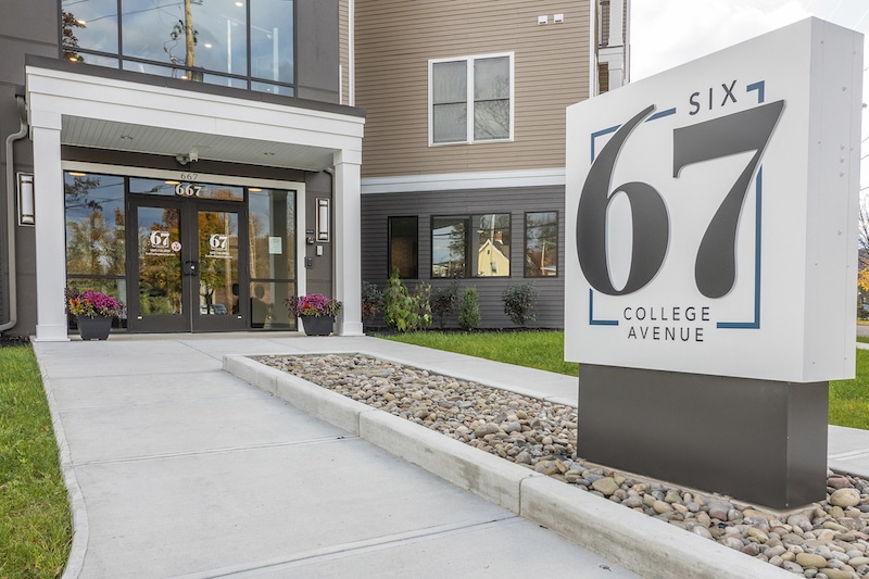 Entrance to a modern building with a sign that reads "67 College Avenue" situated on a landscaped walkway.