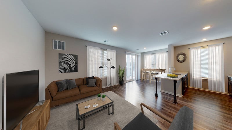 Modern living room and kitchen with hardwood floors, brown sofa, wall art, flat-screen TV, and breakfast bar with stools. Large windows let in natural light.