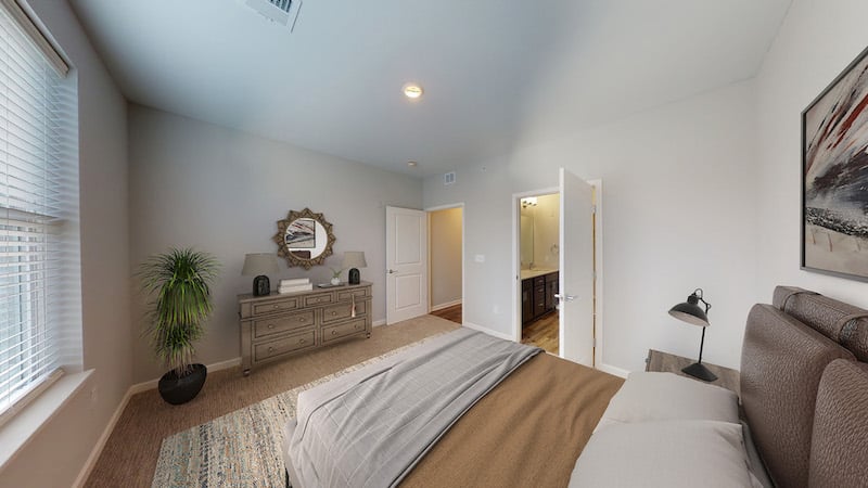 A bedroom with a bed, nightstand, dresser, and a plant. There's a door leading to a bathroom and a window with blinds on the opposite wall.