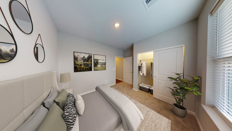 Modern bedroom with a neatly made bed, two landscape paintings, wall mirrors, a potted plant, and an open closet in the corner. Natural light from a window brightens the space.