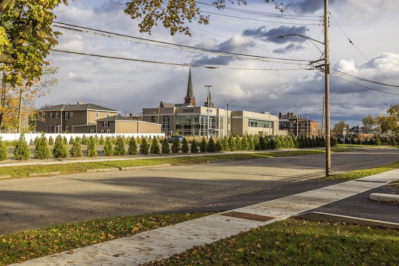 A quiet suburban street with modern buildings in the background and a row of small trees lining the road. A church steeple is visible against a partly cloudy sky.