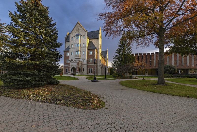 A historic building with a tall gabled roof stands on a paved walkway, surrounded by trees with autumn foliage. Another modern building is visible in the background.