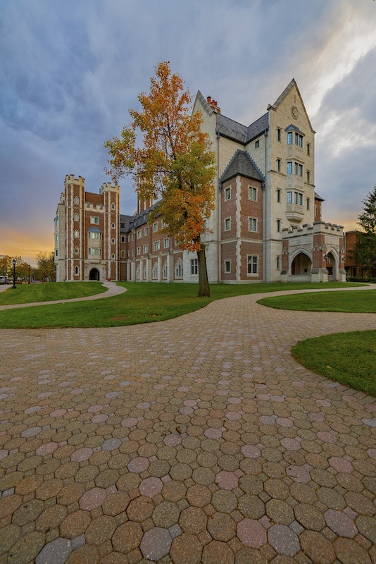 A large, multi-story brick and stone building with peaked roofs stands on a grassy area. A tree with autumn leaves is in front, and a paved walkway leads to the entrance.