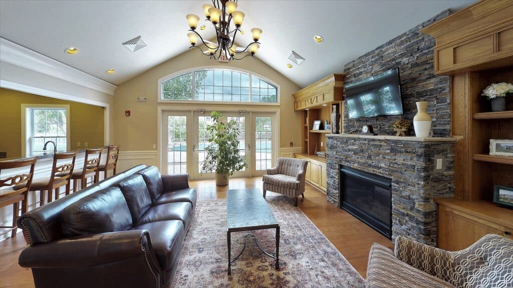 A cozy living room features a leather sofa, stone fireplace, wall-mounted TV, and a wooden dining area with a chandelier. Large windows and glass doors let in natural light.